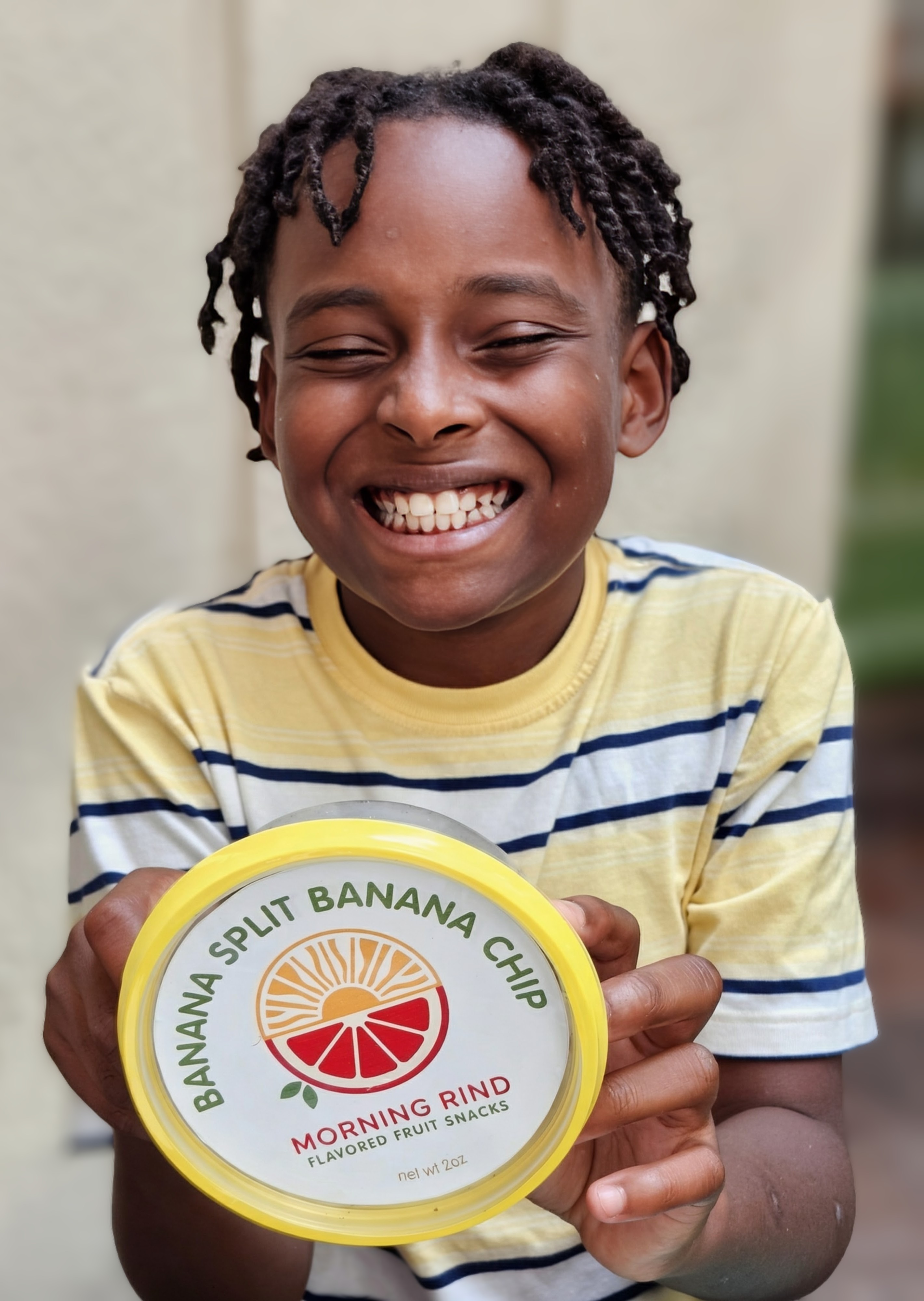 A smiling kid holding a container of Morning Rind Banana Split Banana Chip fruit snacks
